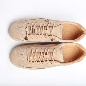 May include: Beige suede trainers with matching laces and metal embellishments. The shoes have a rounded toe and a low profile, suitable for casual wear. The brand name "ANGIE" is visible on the insole. The trainers are a neutral colour.