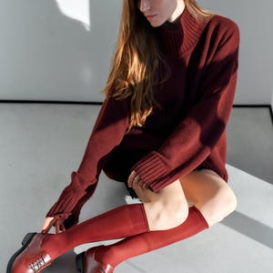 Peut inclure: Une personne porte un pull &agrave; col roul&eacute; bordeaux, des chaussettes montantes et des chaussures Oxford. Le pull a des manches longues et une texture c&ocirc;tel&eacute;e. Les chaussettes et les chaussures sont de la m&ecirc;me nuance de bordeaux. La personne est assise sur une surface gris clair.