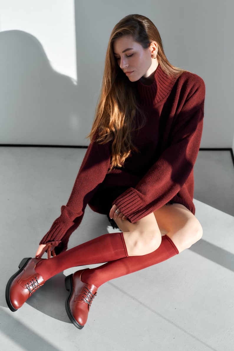Peut inclure: Une personne porte un pull &agrave; col roul&eacute; bordeaux, des chaussettes montantes et des chaussures Oxford. Le pull a des manches longues et une texture c&ocirc;tel&eacute;e. Les chaussettes et les chaussures sont de la m&ecirc;me nuance de bordeaux. La personne est assise sur une surface gris clair.