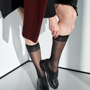 May include: Black leather oxford shoes with a low heel. The shoes are paired with sheer black knee-high stockings. A burgundy sweater sleeve is visible in the upper left corner. The background is a minimalist white and gray.