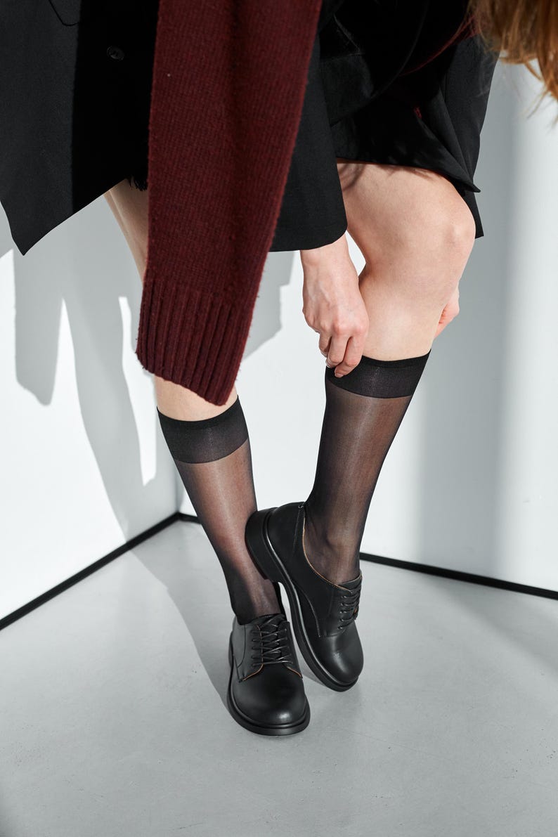 May include: Black leather oxford shoes with a low heel. The shoes are paired with sheer black knee-high stockings. A burgundy sweater sleeve is visible in the upper left corner. The background is a minimalist white and gray.