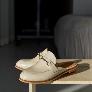 May include: A pair of cream-colored leather mules with a gold-tone buckle detail. The shoes have a rounded toe and a low heel. The mules are displayed on a light-colored surface, showcasing a minimalist design.
