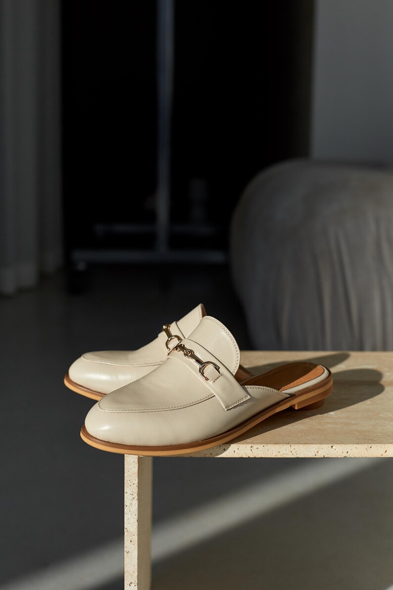 May include: A pair of cream-colored leather mules with a gold-tone buckle detail. The shoes have a rounded toe and a low heel. The mules are displayed on a light-colored surface, showcasing a minimalist design.