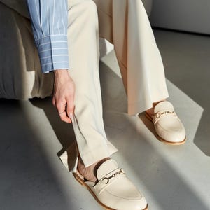 May include: Cream-colored leather slip-on loafers with gold-colored metal accents. The shoes have a rounded toe and a low heel. The image also shows beige pants and a light blue and white striped shirt.