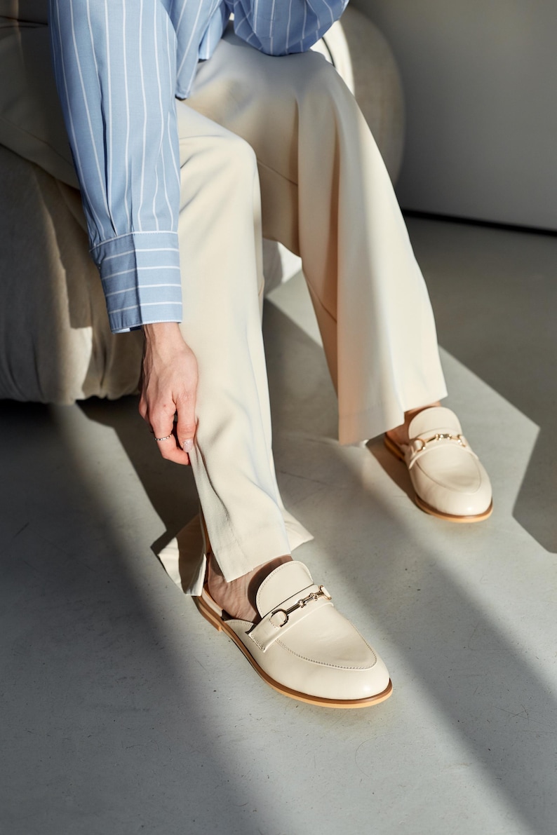 May include: Cream-colored leather slip-on loafers with gold-colored metal accents. The shoes have a rounded toe and a low heel. The image also shows beige pants and a light blue and white striped shirt.