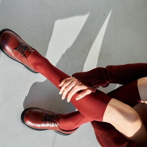 Peut inclure: Chaussures en cuir bordeaux avec lacets et semelles noires. Les chaussures sont associ&eacute;es &agrave; des chaussettes bordeaux assorties qui montent jusqu'&agrave; la cuisse. La personne porte &eacute;galement un pull bordeaux.