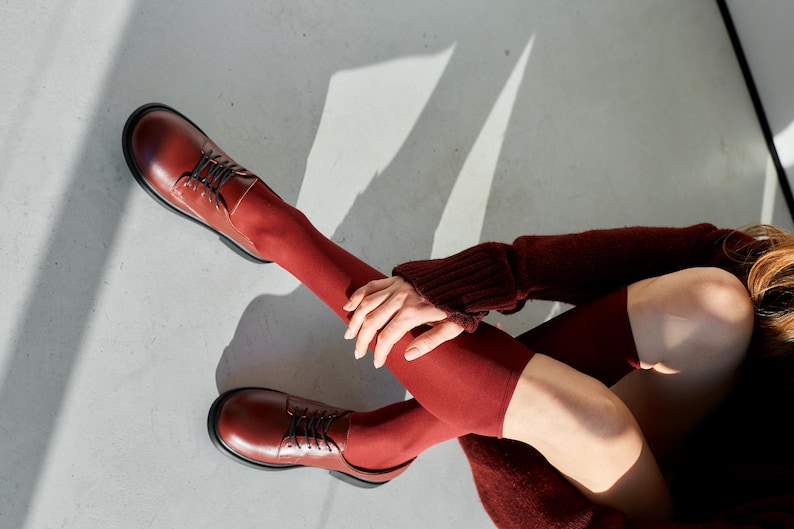 Peut inclure: Chaussures en cuir bordeaux avec lacets et semelles noires. Les chaussures sont associ&eacute;es &agrave; des chaussettes bordeaux assorties qui montent jusqu'&agrave; la cuisse. La personne porte &eacute;galement un pull bordeaux.