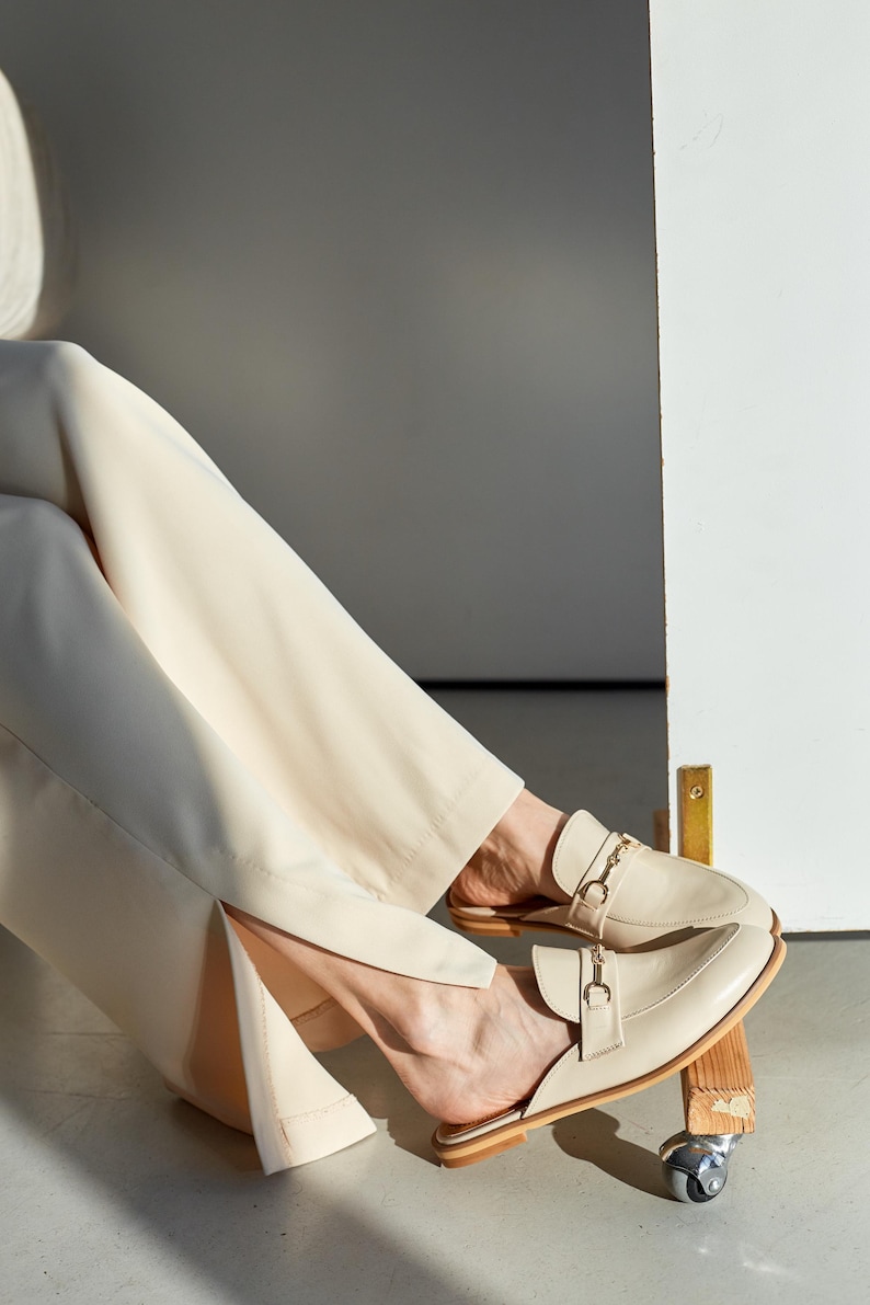May include: Cream-colored leather loafers with gold-tone chain and buckle details. The slip-on shoes have a pointed toe and a low heel. The image shows the shoes on a person's feet, paired with cream-colored pants.