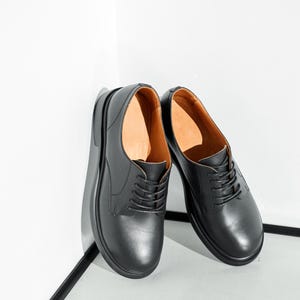 May include: A pair of black leather dress shoes with a rounded toe and a lace-up closure. The shoes have a thick sole and a brown interior. The shoes are leaning against a white wall.