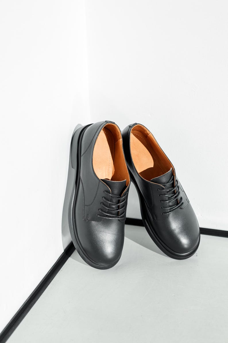May include: A pair of black leather dress shoes with a rounded toe and a lace-up closure. The shoes have a thick sole and a brown interior. The shoes are leaning against a white wall.