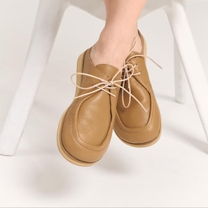 May include: A pair of tan leather shoes with a pebbled texture, featuring a moccasin-style stitched toe box, a thick light-colored sole, and thin beige laces tied in a bow. The shoes have a rounded silhouette and a low-cut ankle design.
