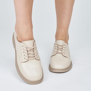 May include: Cream-colored leather shoes with a rounded toe and a thick, light brown sole. The shoes have matching cream-colored laces and a classic Oxford style. The image shows the shoes from a low angle, with the feet of a person wearing them.