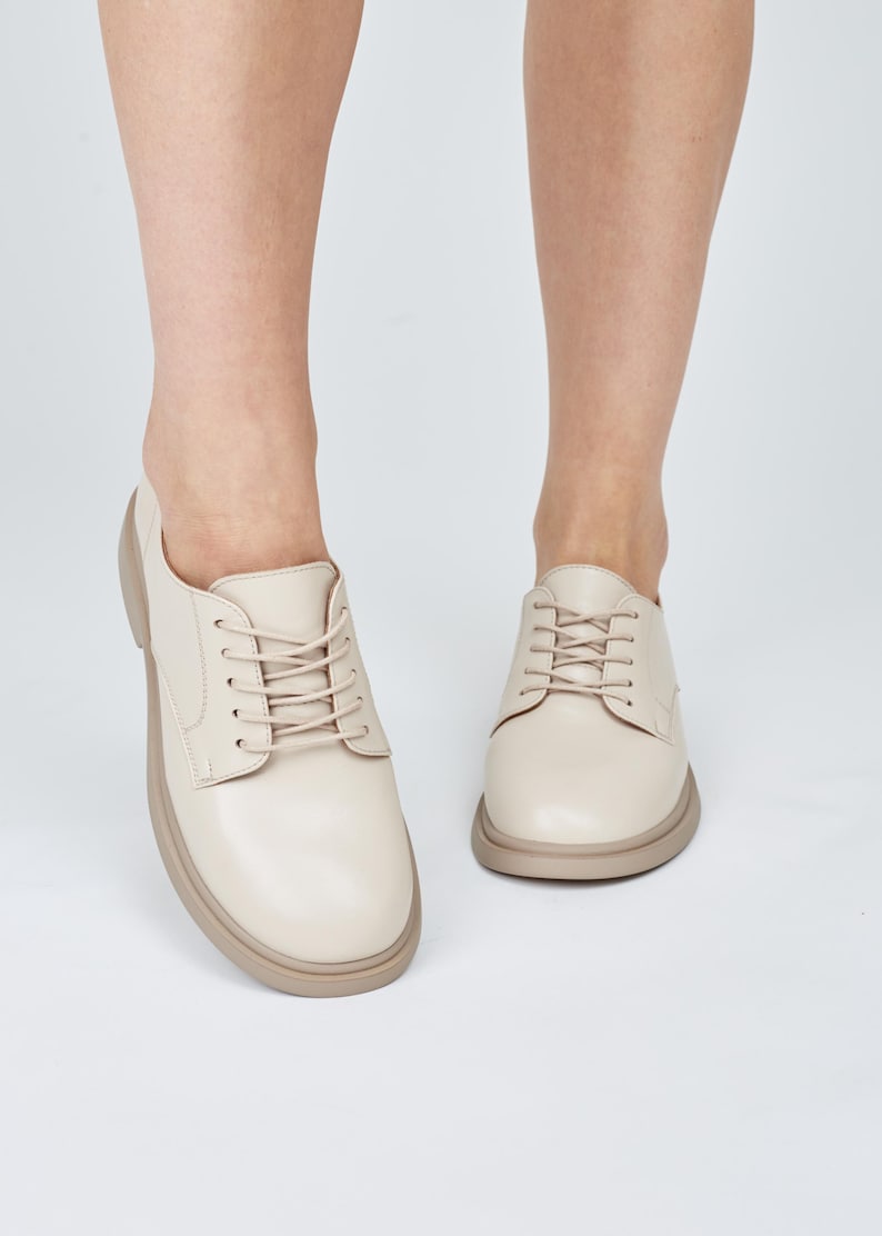 May include: Cream-colored leather shoes with a rounded toe and a thick, light brown sole. The shoes have matching cream-colored laces and a classic Oxford style. The image shows the shoes from a low angle, with the feet of a person wearing them.