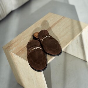 May include: A pair of brown suede slip-on mules with gold-colored metal accents. The shoes have a rounded toe and a low heel. The mules are displayed on a light-colored, textured surface.