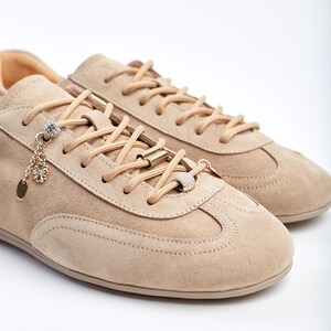 May include: Beige suede trainers with matching laces and decorative gold and crystal accents. The trainers have a classic design with a rounded toe and a low profile, suitable for casual wear.