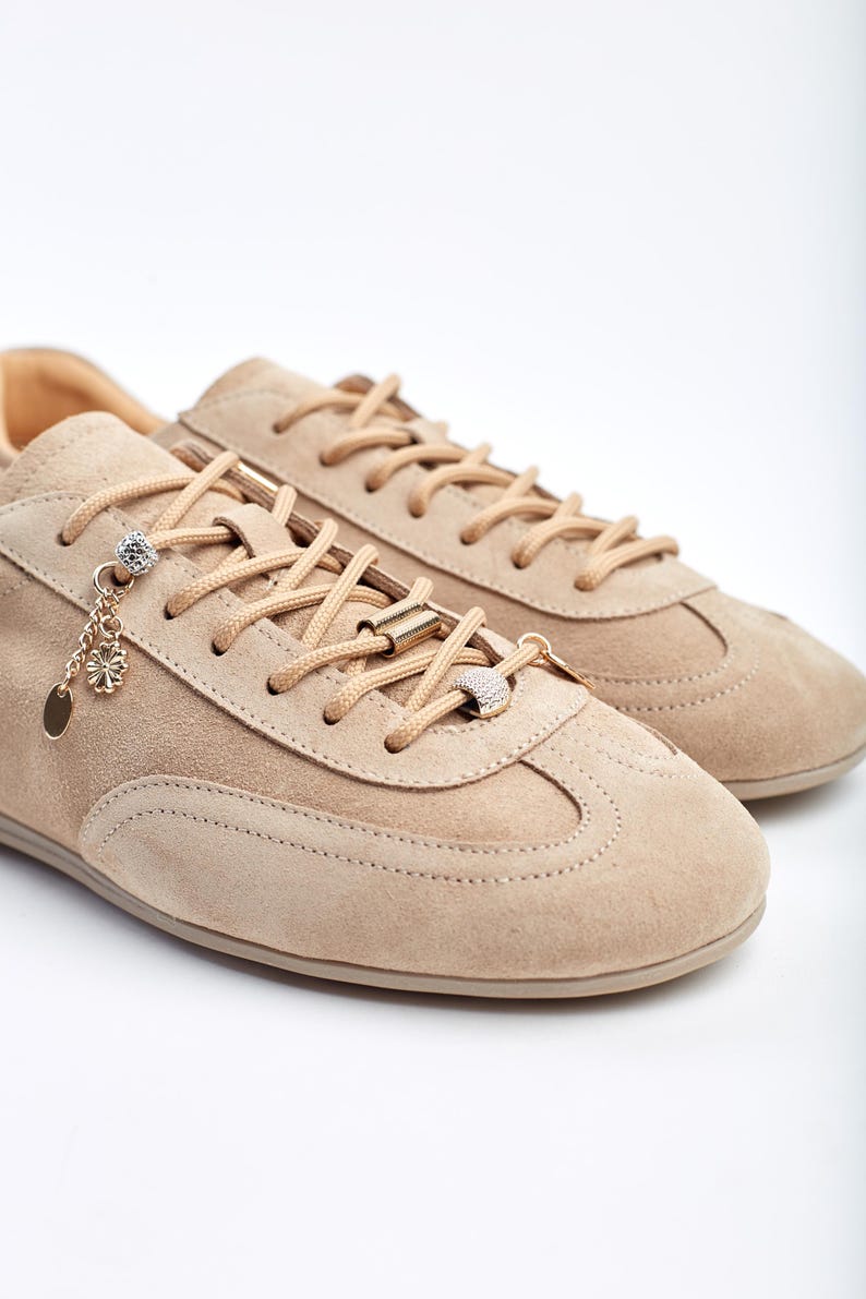 May include: Beige suede trainers with matching laces and decorative gold and crystal accents. The trainers have a classic design with a rounded toe and a low profile, suitable for casual wear.