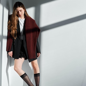 May include: A woman wearing a black blazer, white shirt, and black skirt, paired with black shoes and sheer black knee-high socks. A burgundy knit scarf is draped over the shoulders. The setting is a minimalist white space with shadow accents.