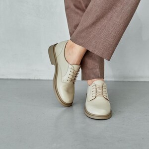 May include: Cream-colored leather Oxford shoes with a rounded toe and a thick, light brown sole. The shoes have matching cream-colored laces and are worn with brown trousers. The shoes are a classic style.
