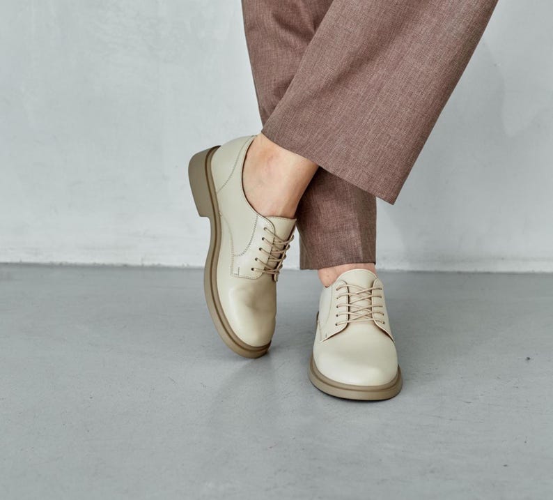 May include: Cream-colored leather Oxford shoes with a rounded toe and a thick, light brown sole. The shoes have matching cream-colored laces and are worn with brown trousers. The shoes are a classic style.