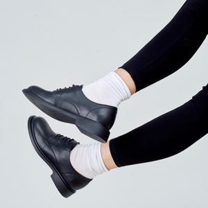 May include: Black leather derby shoes with laces, paired with white socks and black leggings. The shoes have a classic design with a rounded toe and a low heel. The image is set against a plain white background, highlighting the footwear.
