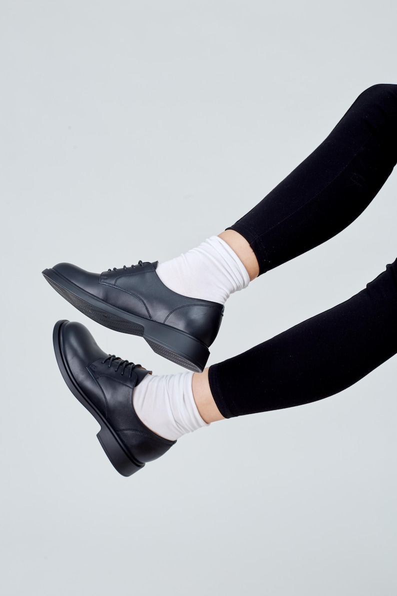 May include: Black leather derby shoes with laces, paired with white socks and black leggings. The shoes have a classic design with a rounded toe and a low heel. The image is set against a plain white background, highlighting the footwear.
