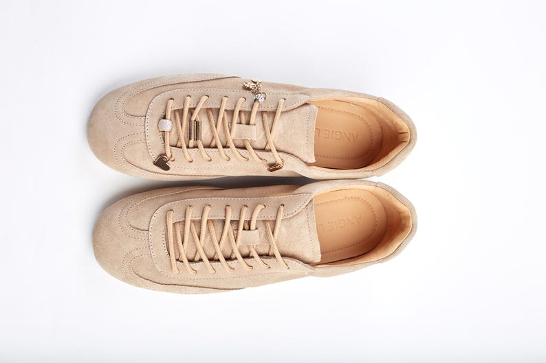 May include: Beige suede trainers with matching laces and metal embellishments. The shoes have a rounded toe and a low profile, suitable for casual wear. The brand name "ANGIE" is visible on the insole. The trainers are a neutral colour.