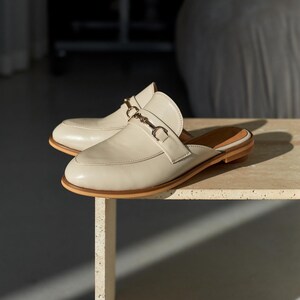 Elegant women’s leather mules in cream beige with a classic slip-on design. Featuring a low wooden heel, rounded toe, and metal horsebit detail across the upper strap, these versatile backless loafers combine comfort with timeless style. Perfect for casual wear, office outfits, or chic everyday looks, crafted from smooth genuine leather with a durable sole for long-lasting use.