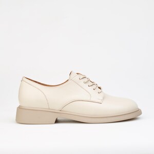 May include: Cream-colored leather oxford shoe with a rounded toe and a low, stacked heel. The shoe features tonal laces and stitching details. The sole is a light beige color.