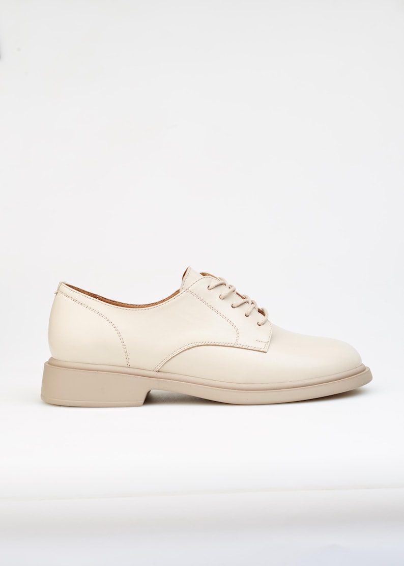 May include: Cream-colored leather oxford shoe with a rounded toe and a low, stacked heel. The shoe features tonal laces and stitching details. The sole is a light beige color.
