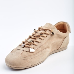 May include: A beige suede trainer with decorative charms on the laces. The charms include a heart, a diamond, and a ring. The shoe has a rounded toe and a low profile sole. The laces are beige, matching the shoe's colour.