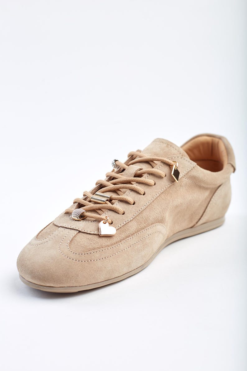 May include: A beige suede trainer with decorative charms on the laces. The charms include a heart, a diamond, and a ring. The shoe has a rounded toe and a low profile sole. The laces are beige, matching the shoe's colour.