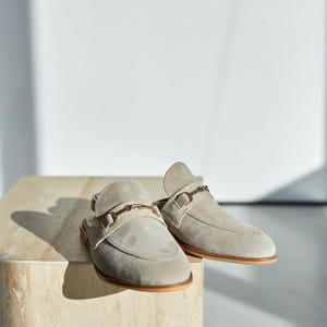 May include: A pair of light gray suede slip-on loafers with a gold-colored metal buckle detail. The shoes have a rounded toe and a brown sole. The footwear is displayed on a light-colored, textured surface.