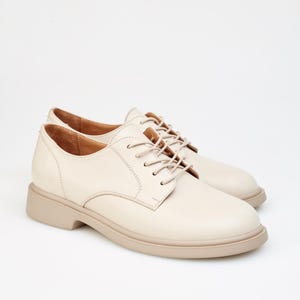 May include: A pair of cream-colored leather Oxford shoes with matching laces. The shoes have a rounded toe and a thick, beige sole. The style is classic and versatile, suitable for various occasions.