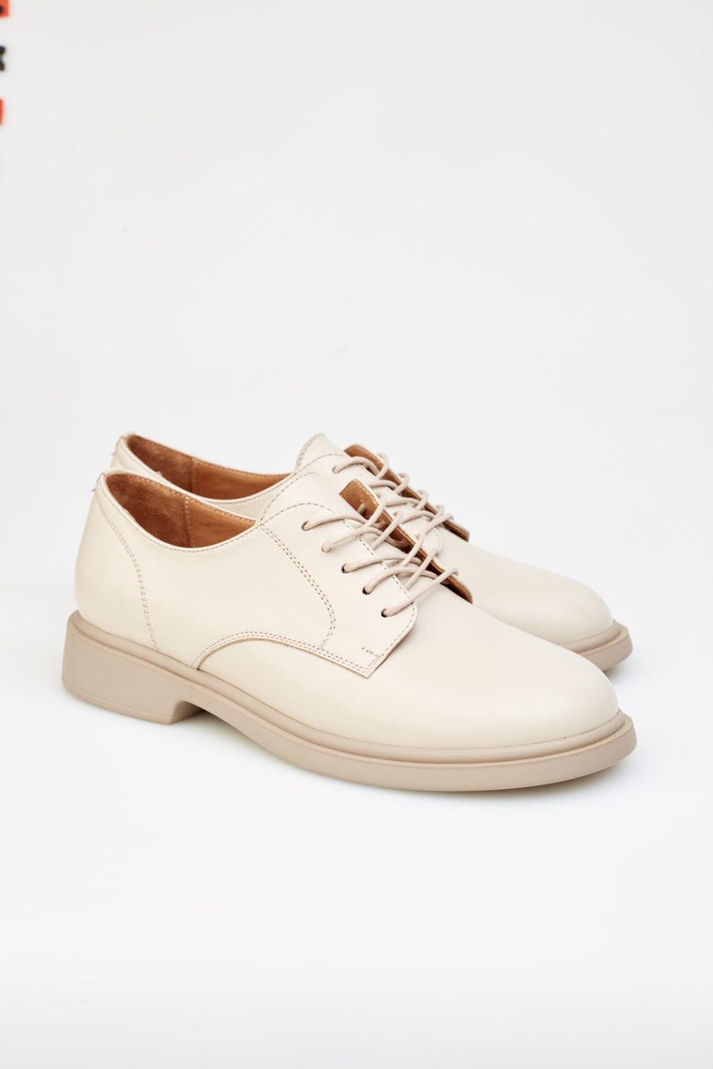 May include: A pair of cream-colored leather Oxford shoes with matching laces. The shoes have a rounded toe and a thick, beige sole. The style is classic and versatile, suitable for various occasions.