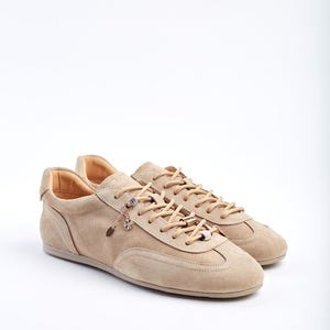May include: A pair of beige suede trainers with matching laces. The shoes have a low profile and a decorative charm hanging from the laces. The trainers are set against a white background.