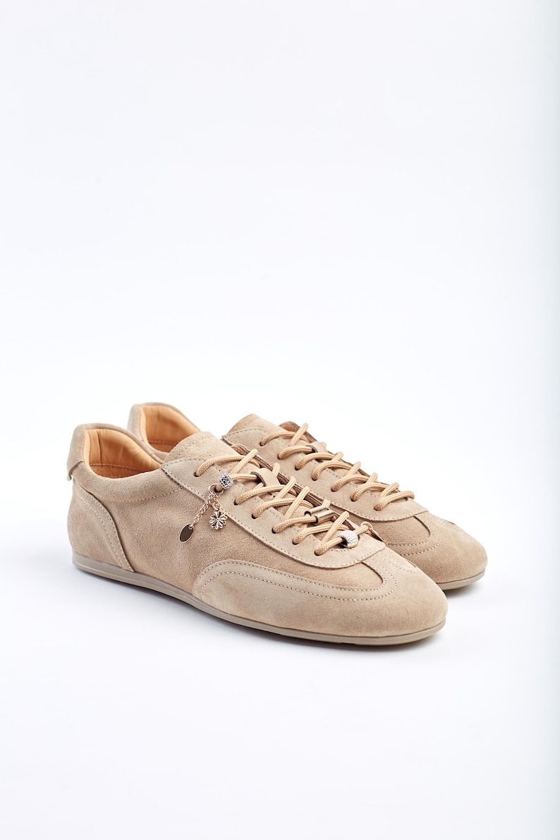 May include: A pair of beige suede trainers with matching laces. The shoes have a low profile and a decorative charm hanging from the laces. The trainers are set against a white background.