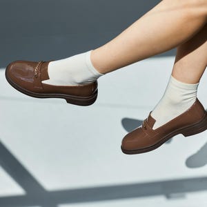 May include: Brown patent leather loafers with gold-colored chain detail. The shoes are worn with white socks. The loafers have a classic design with a rounded toe and a low heel. The shoes are a rich, warm brown color.