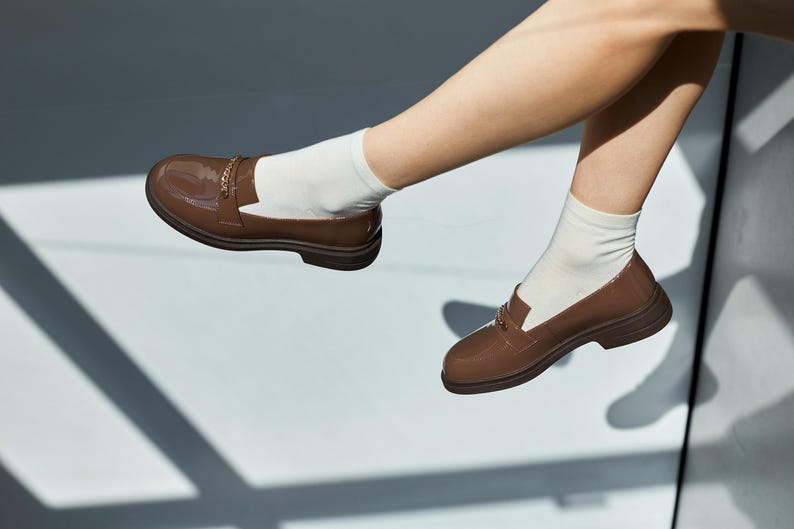 May include: Brown patent leather loafers with gold-colored chain detail. The shoes are worn with white socks. The loafers have a classic design with a rounded toe and a low heel. The shoes are a rich, warm brown color.