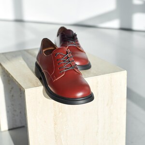 Peut inclure: Paire de chaussures de ville en cuir bordeaux avec lacets et semelles noires. Les chaussures sont pr&eacute;sent&eacute;es sur un socle carr&eacute; de couleur claire sur fond blanc. Les chaussures ont un aspect classique et poli.