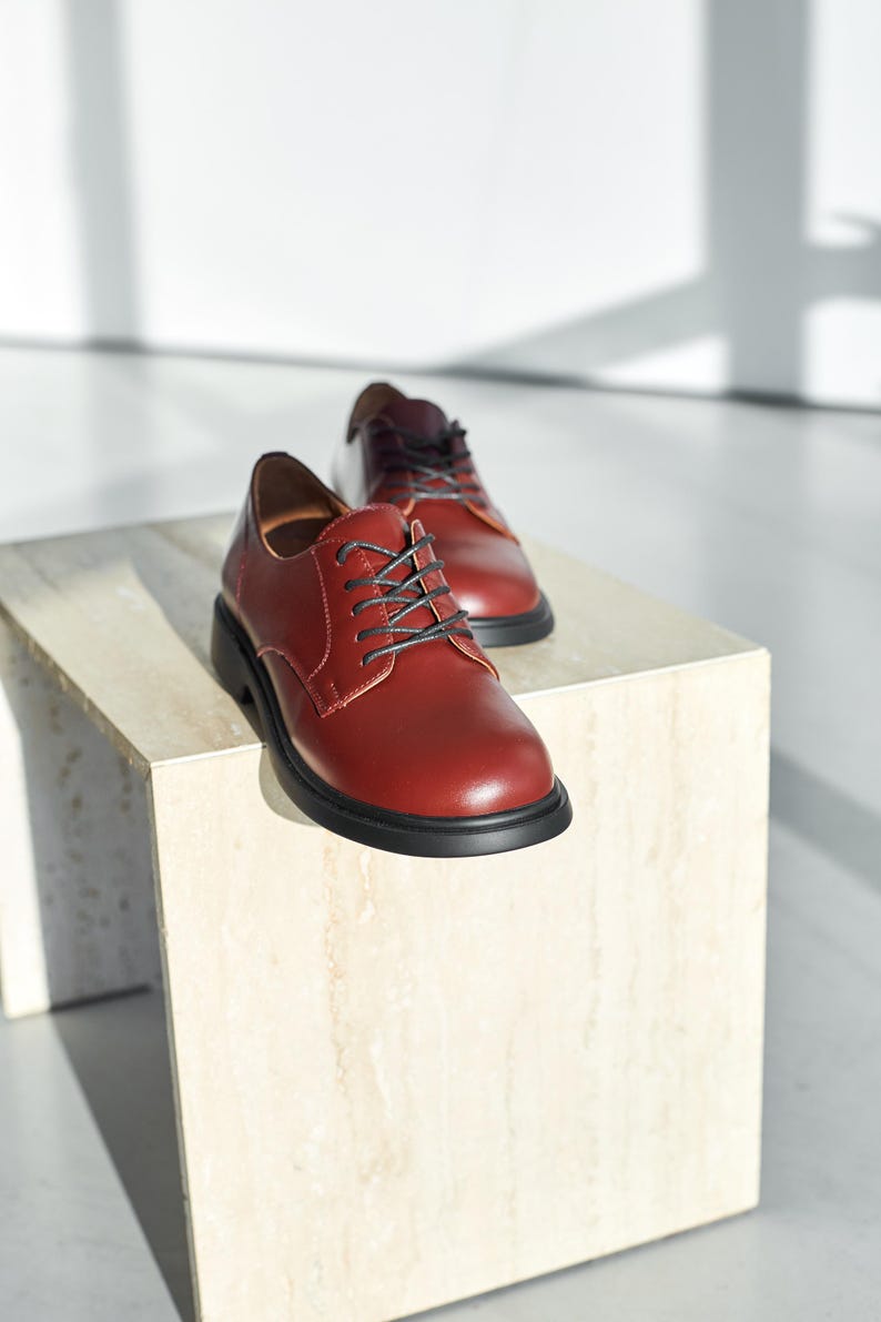 Peut inclure: Paire de chaussures de ville en cuir bordeaux avec lacets et semelles noires. Les chaussures sont pr&eacute;sent&eacute;es sur un socle carr&eacute; de couleur claire sur fond blanc. Les chaussures ont un aspect classique et poli.