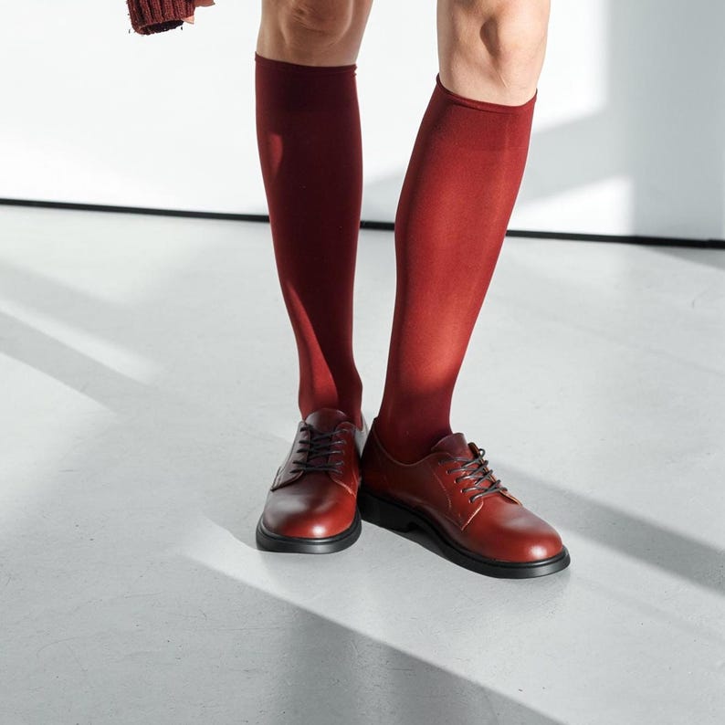 Peut inclure: Chaussettes hautes bordeaux et chaussures en cuir marron avec semelles et lacets noirs. Les chaussures sont de style Oxford classique. L'ensemble est pos&eacute; sur une surface blanche.