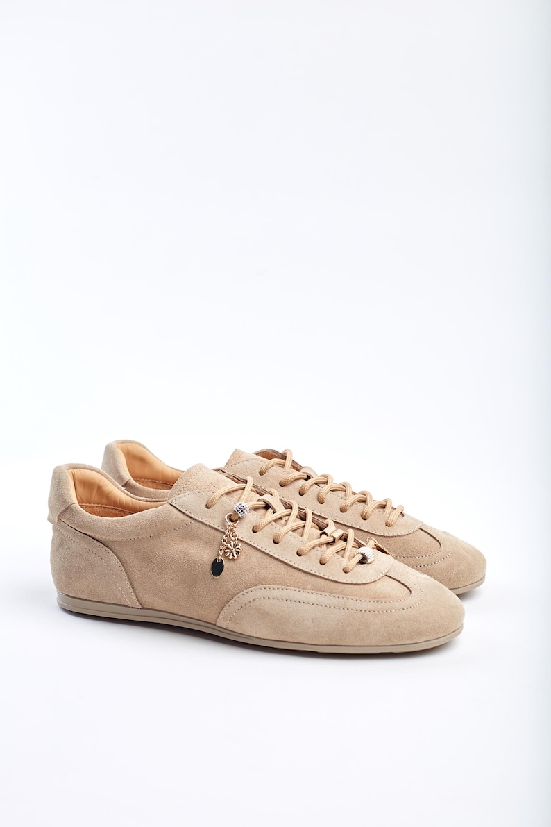 Low-top women’s sneakers in soft beige suede, with a streamlined silhouette and tonal laces threaded through metal eyelets and decorative charm accents. The supple suede upper features subtle stitching lines and curved paneling for a retro-inspired yet modern look. A cushioned leather-lined interior and slim rubber sole provide all-day comfort and traction, making these versatile trainers ideal for casual weekend wear or stylish everyday outfits.