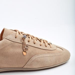 May include: A close-up of a beige suede trainer with matching laces. The shoe has decorative charms hanging from the laces, including a small flower and a circular pendant. The trainer is set against a white background.