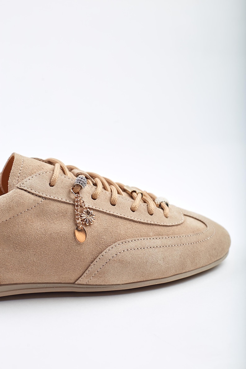 May include: A close-up of a beige suede trainer with matching laces. The shoe has decorative charms hanging from the laces, including a small flower and a circular pendant. The trainer is set against a white background.