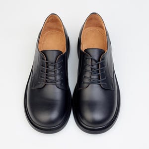 May include: A pair of black leather Oxford shoes with a rounded toe and a lace-up closure. The shoes have a thick sole and a light brown interior. The shoes are displayed on a white background.