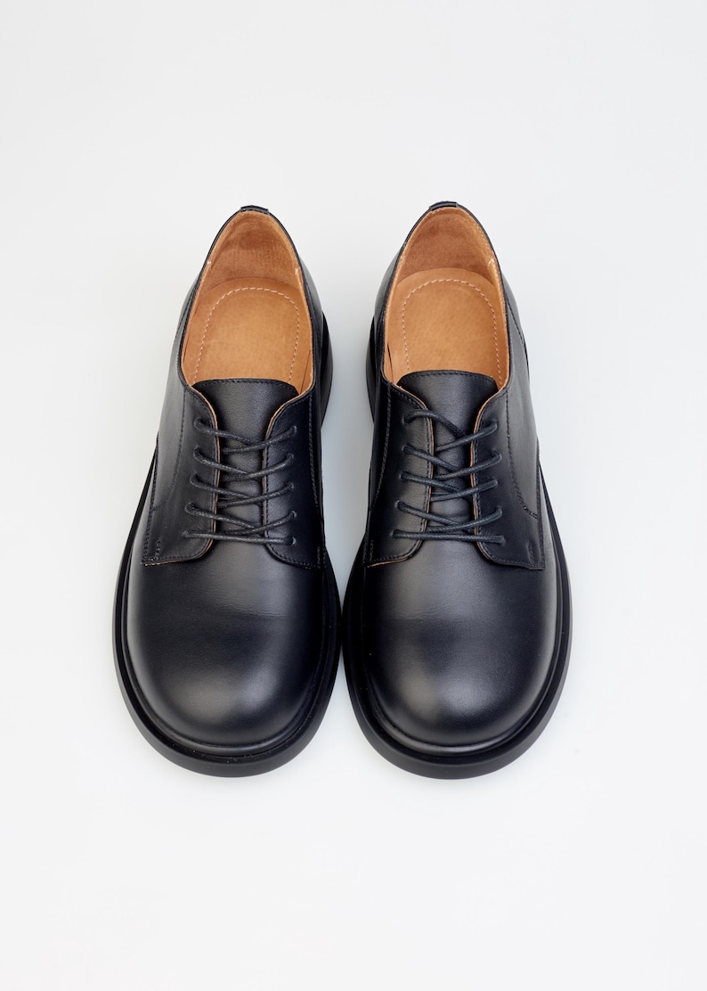 May include: A pair of black leather Oxford shoes with a rounded toe and a lace-up closure. The shoes have a thick sole and a light brown interior. The shoes are displayed on a white background.