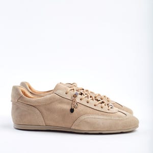 May include: Beige suede trainers with a low profile and lace-up design. The shoes have decorative charms on the laces. The neutral colour palette makes them suitable for casual wear. The trainers are made of suede.