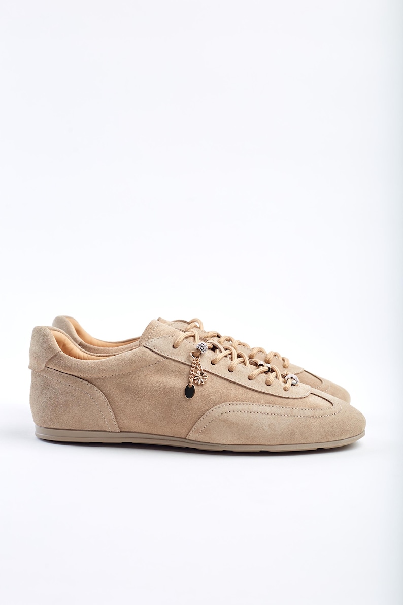 May include: Beige suede trainers with a low profile and lace-up design. The shoes have decorative charms on the laces. The neutral colour palette makes them suitable for casual wear. The trainers are made of suede.
