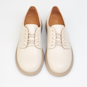 May include: A pair of cream-colored, lace-up shoes with a rounded toe and a thick, light brown sole. The shoes have a classic oxford style and are set against a white background.