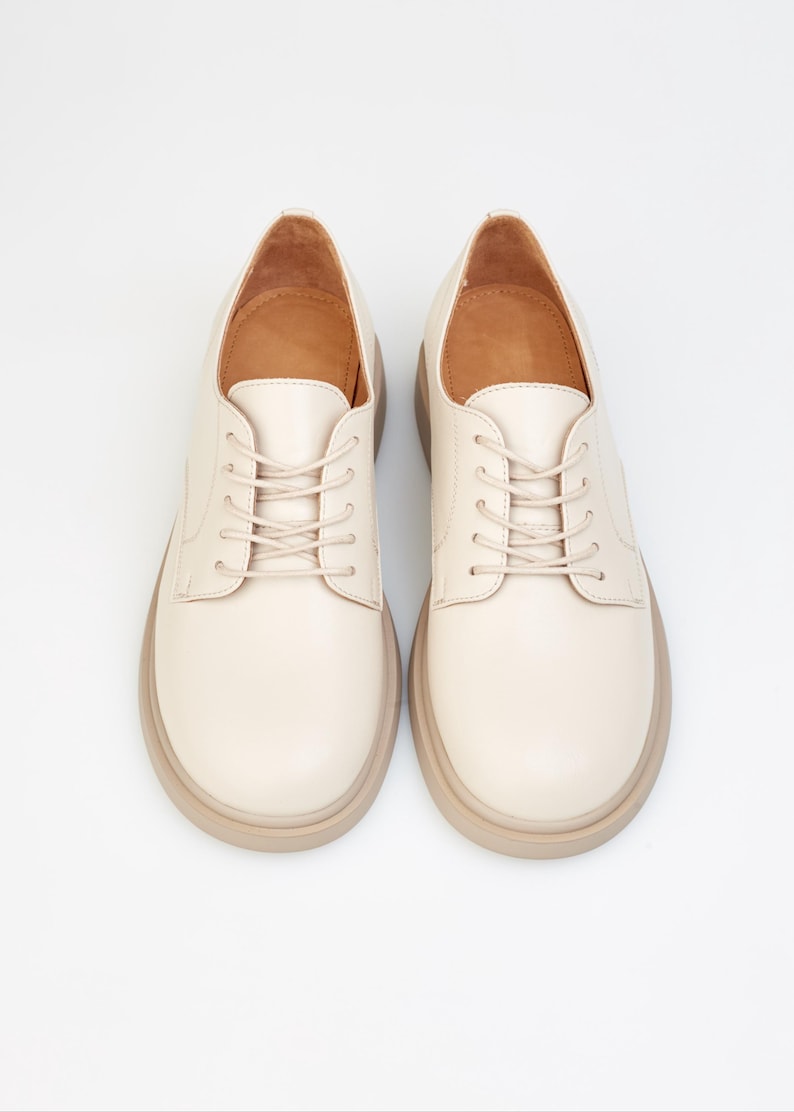 May include: A pair of cream-colored, lace-up shoes with a rounded toe and a thick, light brown sole. The shoes have a classic oxford style and are set against a white background.